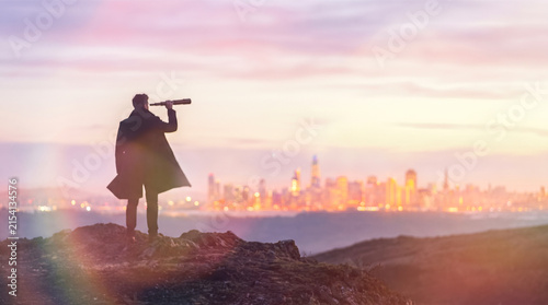 Male silhouette standing on rocky hill looking through vintage telescope toward glowing distant city skyline at dawn representing business forecasting and strategic planning
