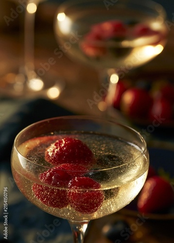 Two glasses of champagne on a table. the glasses are filled with a golden-colored liquid, likely champagne, and are garnished with fresh raspberries.