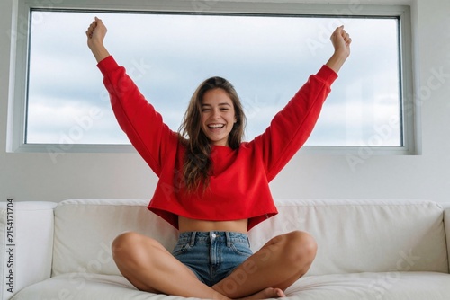 Young woman celebrates on sofa by large window. She sits cross-legged with arms raised in excitement. Cheerful lady feels joy and happiness at home.