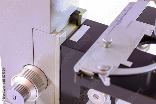 Detailed view of a laboratory microscope stage showing a metal scale, specimen holder and adjustment controls for precise sample positioning.