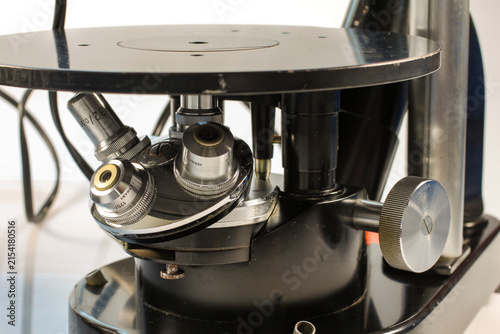 Close-up of a vintage laboratory microscope showing the rotating objective turret, multiple objective lenses, mechanical focusing controls and specimen stage, isolated on a light background.