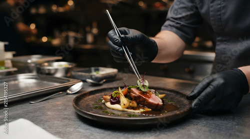 Professional chef in black gloves carefully plating a gourmet dish with culinary tweezers in a restaurant kitchen