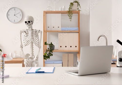Interior of medical office with human skeleton model, shelf unit and workplace