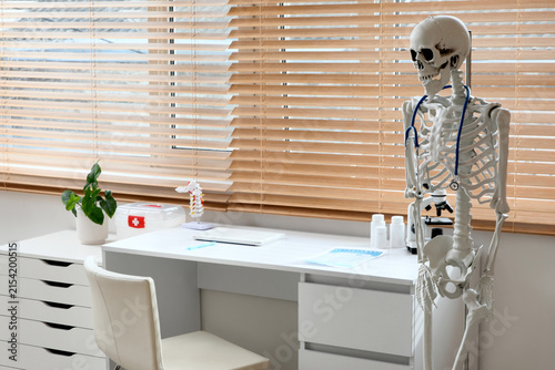 Human skeleton model with stethoscope near desk in medical office interior