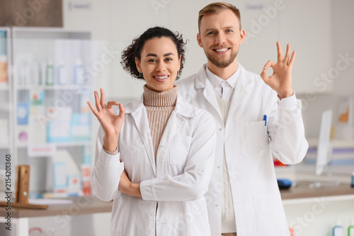 Smiling male and female pharmacists in white coats making an ok gesture in a ...