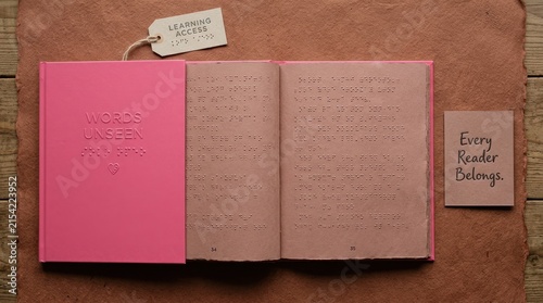 Pink braille book titled Words Unseen lies open on textured paper next to learning access tags and an every reader belongs card, promoting inclusive education, accessibility for visually impaired