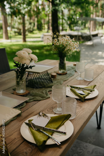 A table set with plates, forks, spoons, napkins, and flowers in transparent vases in an open space for a wedding, anniversary, or birthday celebration