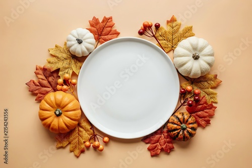 White ceramic plate centered on a beige background, surrounded by a decorative wreath of autumn maple leaves, small pumpkins, and red berries, seasonal thanksgiving or holiday dining