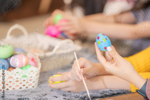 Fotografie Above a wicker basket heaped with bright Easter eggs, small hands and an adult hand steadying a hand-painted Easter egg above a paint palette on a happy holiday morning