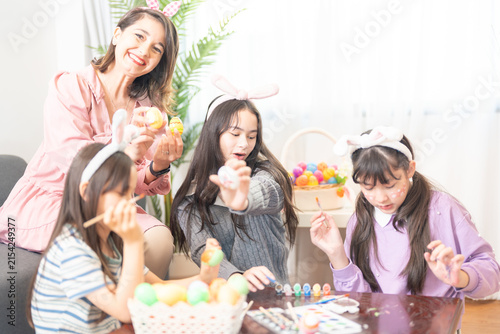 Fotografie A young mother and two little girls painting Easter eggs together with fine brushes at a wooden coffee table strewn with brushes, paints and eggs on a happy holiday morning