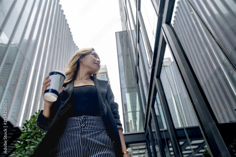 Fototapeta premium Asian businesswoman walks briskly with mouth slightly open and gaze lifted up and right while holding coffee beneath Bangkok's towers, on a busy sidewalk beneath Bangkok's glass skyline.