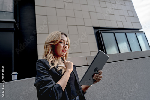Canvastavla An Asian businesswoman with long blonde wavy hair stands beside a stone-clad tower, coffee cup in hand, touching her chin thoughtfully while glancing at her dark tablet outdoors