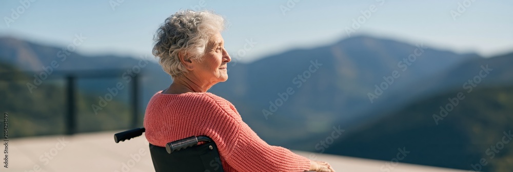 Fototapeta premium Elderly woman in wheelchair facing panoramic mountain landscape background for senior care poster and wellness banner design