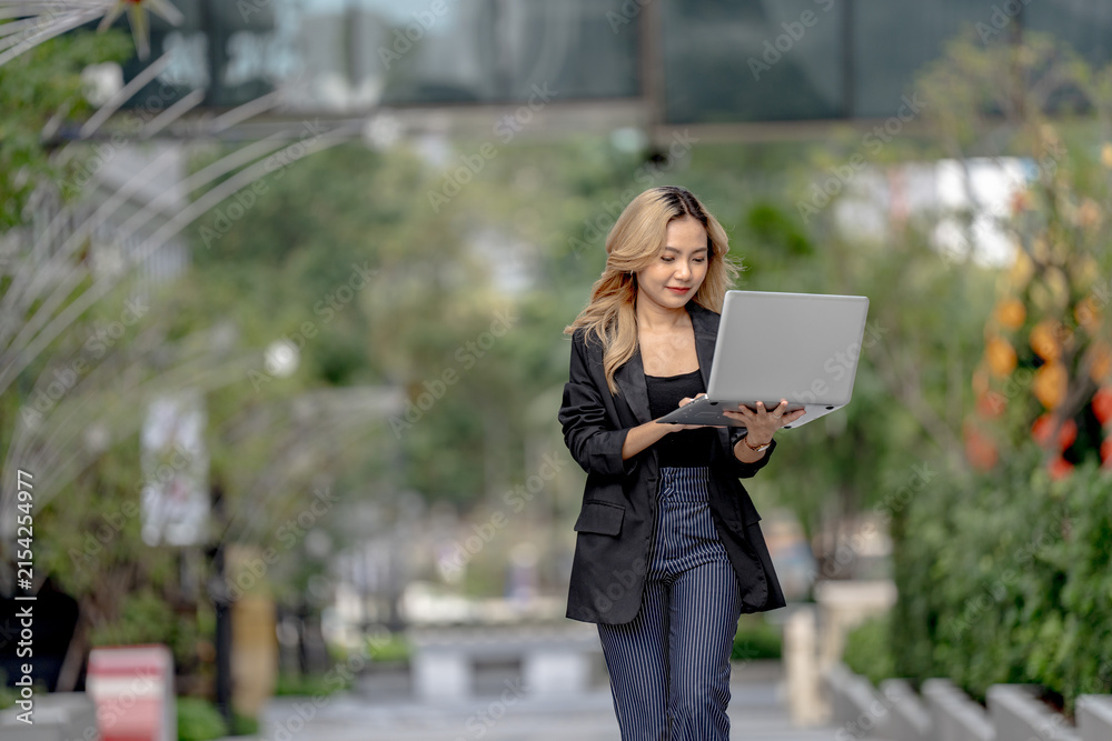 Fototapeta premium Asian businesswoman with long blonde wavy hair strides along a landscaped garden path in Bangkok's business district, holding an open laptop and glancing down toward its glowing screen.