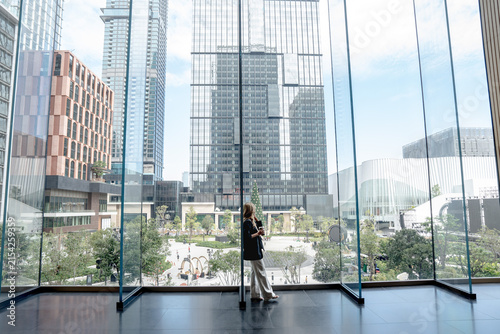 Fotografie Businesswoman leans slightly while talking on her phone near the glass wall, her weight shifted onto one leg, the Bangkok plaza and skyline stretching out beyond
