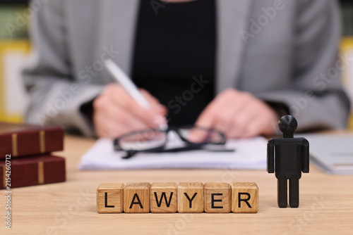 At a tidy desk, a focused lawyer prepares legal documents while a playful wooden block spells out lawyer. Glasses and notepad complete the scene, showcasing professionalism.