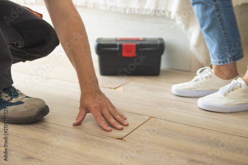 Handyman carefully examining damaged laminate floorboards in a domestic setting. Preparing for floor repair or renovation work while a homeowner stands by