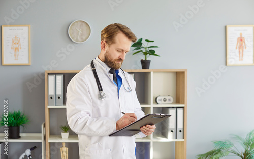 Male physician or general practitioner writing down notes, holding clipboard, posing in medical office, has serious expression, wearing white coat with stethoscope, preparing for appointment