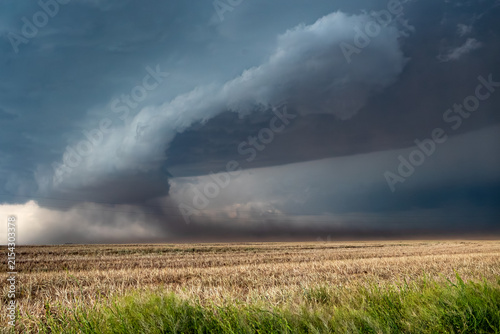 View of a powerful supercell storm with dark, rotating clouds and a shelf cloud hanging over a vast, dry agricultural field under a dramatic, stormy sky.