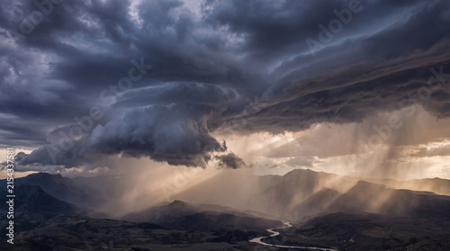 Epic storm clouds gather over majestic mountains with sun rays breaking through rain shafts at a dramatic sunset.