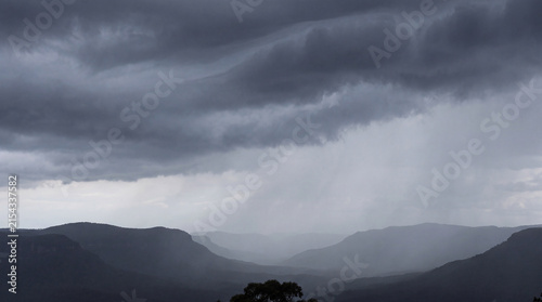 Dramatic rain and dark storm clouds over a majestic mountain range, creating a misty, atmospheric landscape.