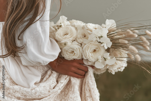 Boho bride from behind holding ivory ranunculus and rose bouquet , lace wedding gown and flowing hair in soft natural light.