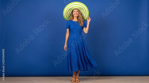 Smiling woman in blue dress and large striped sun hat on blue background.