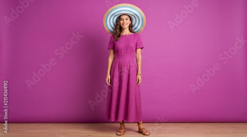 Stylish woman in purple maxi dress wearing striped sun hat against magenta wall.