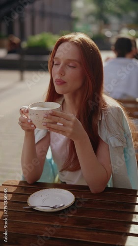 Charming young redhead woman savoring warm cup of coffee sitting alone at wooden table in summer street cafe. Vertical portrait of pretty lady enjoying peaceful moment during sunny day in city.