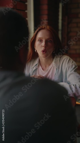 Serious young woman with red hair sitting at table in cafe, gesturing and talking with man during intense and emotional conversation, sharing perspective, vertical shot, rear back view, slow motion.