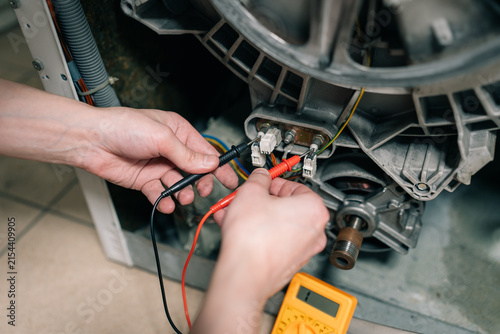 Hands of a professional technician using a digital yellow multimeter to test electrical contacts and voltage in a broken washing machine.	
