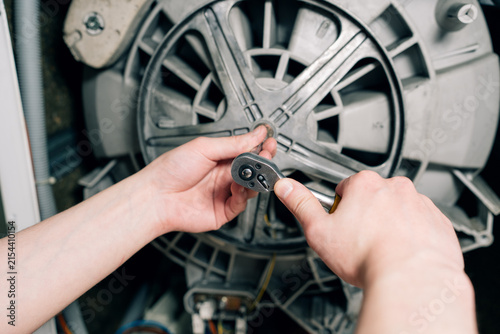 Hands of a mechanic using a socket ratchet wrench tool to tighten or unscrew a bolt on a washing machine drum pulley.	