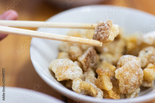 Chopsticks holding crispy deep fried pork rind over white bowl in Asian restaurant.