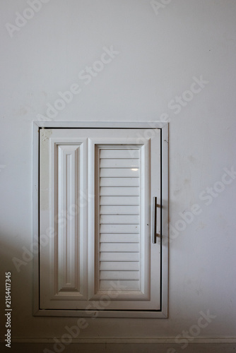 Small white louvered plastic door mounted on white wall for utility access in building interiors.