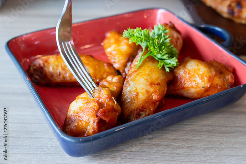 Fork stabbing roasted barbecue chicken wing in red ceramic dish on wooden table.