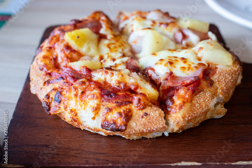 Small Hawaiian pizza topped with ham pineapple chunks and melted cheese on wooden tray.