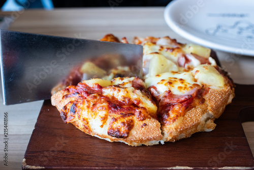 Stainless steel spatula lifting a slice of Hawaiian pizza with pineapple ham and melted cheese on wooden board.