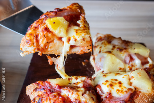 Lifting a slice of Hawaiian pizza with melted cheese pull and pineapple toppings using metal spatula.