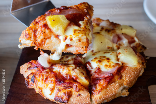 Lifting a slice of Hawaiian pizza with melted cheese pull and pineapple toppings using metal spatula.
