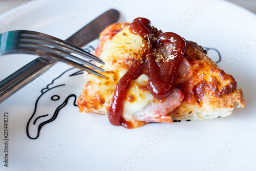 Hawaiian pizza slice drizzled with tomato ketchup and oregano served on white plate with fork and knife.