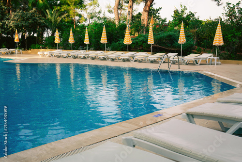 Outdoor swimming pool with blue water, empty sun loungers and yellow striped umbrellas. Luxury resort hotel relaxation zone, summer vacation concept.	
