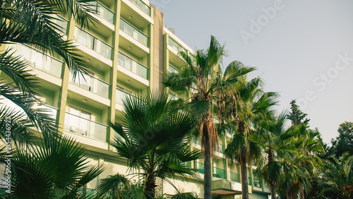 Modern hotel building facade with balconies behind lush green palm trees. Summer vacation resort concept, tropical holiday accommodation.	
