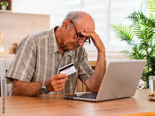 Worried senior man reviewing his bank account and credit card online while sitting at his home. He is feeling stressed and confused about debt, expenses and household finances.