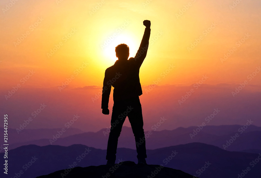 Fototapeta premium Silhouette of a triumphant person standing on a mountain peak at sunset with raised fist