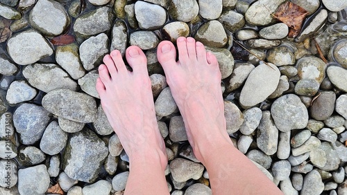Top-down view of bare human feet on a bed of dry, smooth pebbles and rounded stones. Neutral earthy tones and organic shapes create a natural, minimalist composition