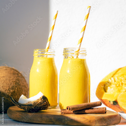Pumpkin and coconut smoothies in glass bottles with striped straws, fresh coconut, mango and cinnamon on a wooden board, closeup