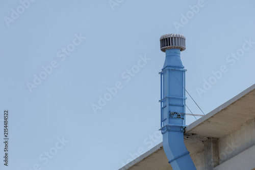 Industrial ventilation duct with turbine vent cap rising above a concrete rooftop against a clear sky.