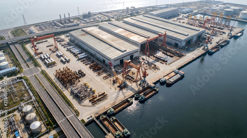 Aerial view of an offshore equipment manufacturing facility with large warehouses, gantry cranes, and cargo barges docked along a concrete pier