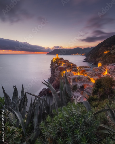 View of the historic coastal village of Vernazza at sunset, with illuminated stone buildings perched on the cliffs overlooking the Mediterranean Sea, Vernazza, Liguria, Italy.