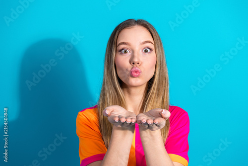 Bild auf Leinwand Young woman blowing kiss at camera against blue background wearing bright pink a